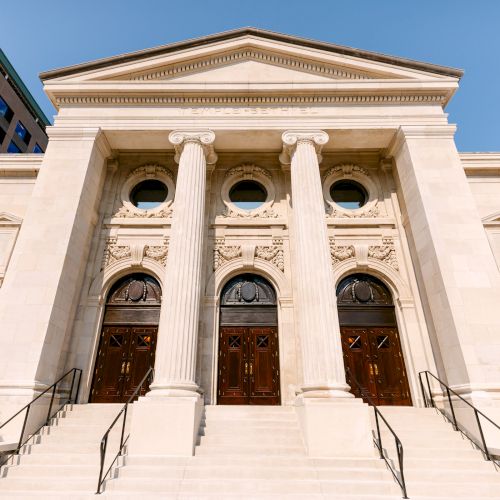 The image shows a neoclassical building with three large wooden doors, pillars, and ornate carvings, likely a religious or civic structure.