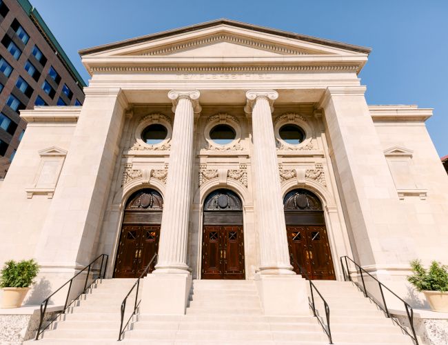 The image shows a neoclassical building with three large wooden doors, pillars, and ornate carvings, likely a religious or civic structure.
